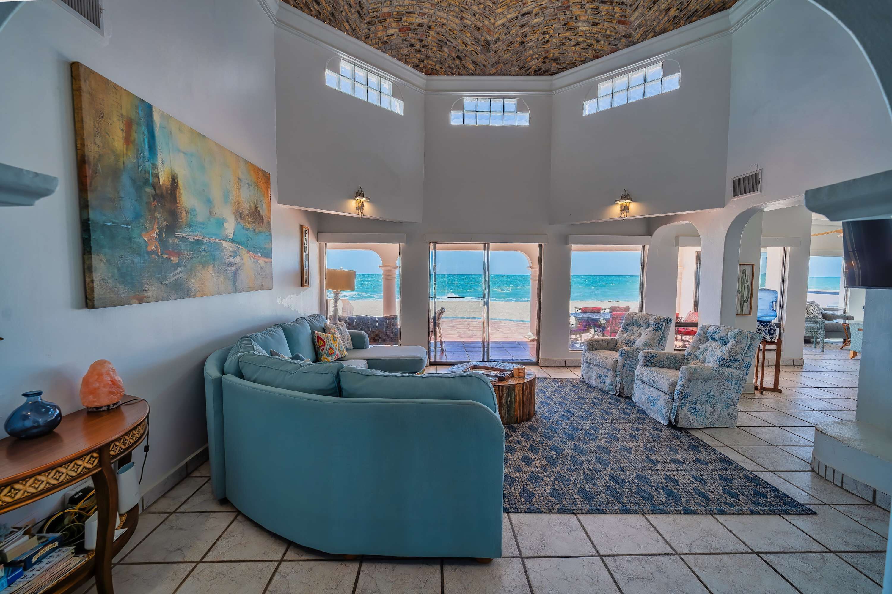 Een ruime woonkamer met een grote bank, fauteuils en uitzicht op het strand door grote ramen.