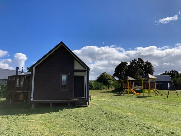 A small, dark-colored building with a gabled roof, situated in a grassy area with a playground and trees in the background.