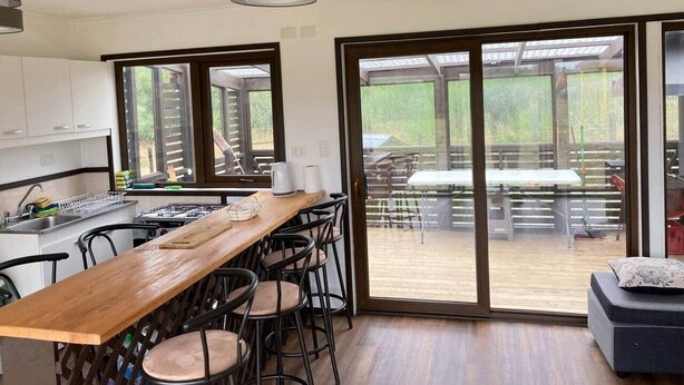 Uma cozinha moderna com bancada de madeira, banquetas e uma porta de vidro deslizante para um pátio externo com mesas e cadeiras.