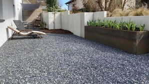 Un patio con fondo in ghiaia, una sedia a sdraio, un vaso con piante verdi e un edificio bianco con tetto rosso.