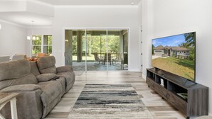 Ein modernes Wohnzimmer mit einer Couch, einem Fernseher und Blick auf ein Haus durch Glastüren.
