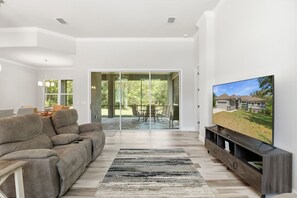 Un salon moderne avec un canapé, un téléviseur et une vue sur une maison, visible à travers des portes vitrées.
