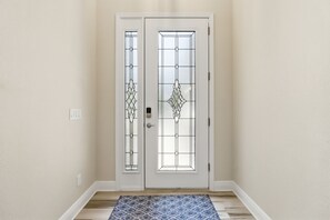 Une porte blanche avec un panneau de verre et un trou de serrure, un tapis à motifs sur le sol et un interrupteur sur le mur.