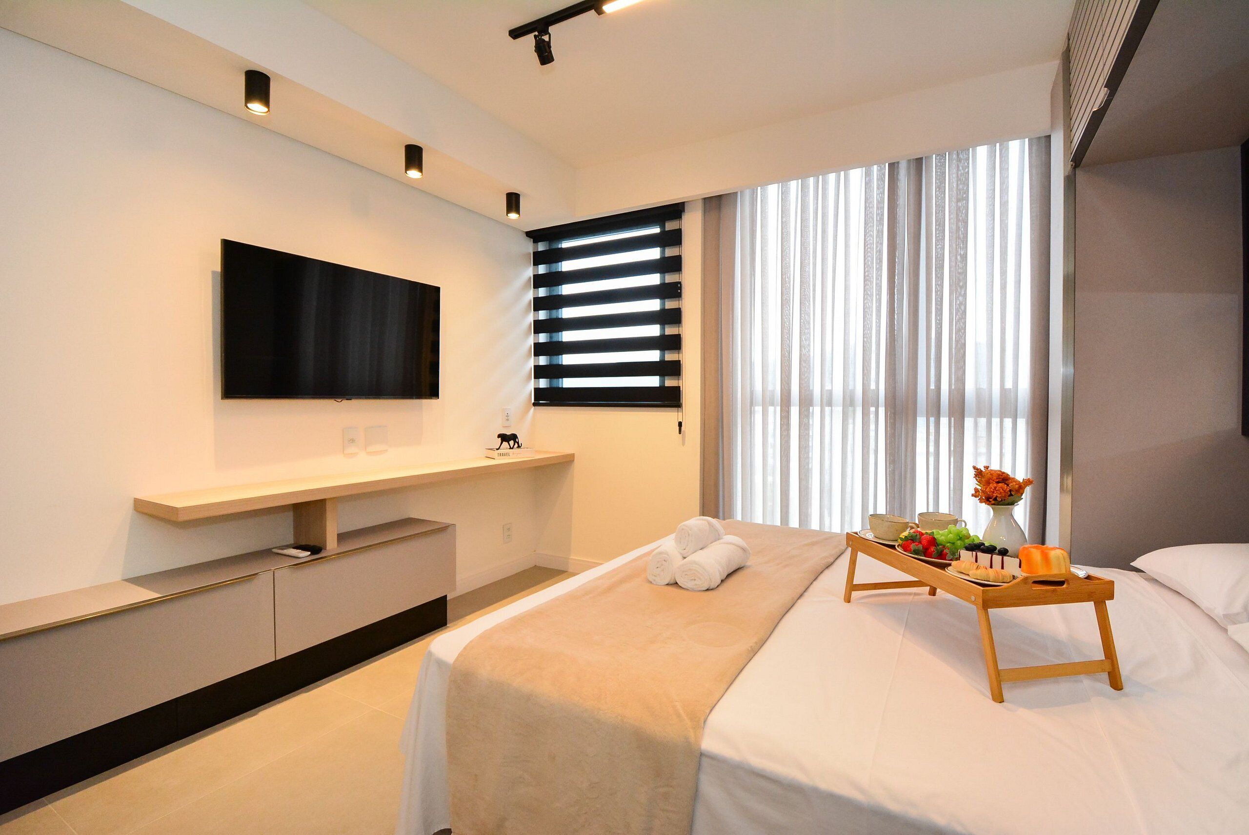 Una habitación de hotel moderna con una televisión de pantalla plana, una mesita de noche de madera con una bandeja de aperitivos, una cama con una manta color beige y un ventanal con persianas.