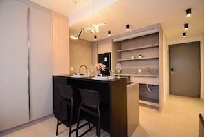 A modern kitchen with a central island, bar stools, a refrigerator, and built-in shelves. - Studio em frente a UFSC e HU - VIVA 207R (Florianópolis)