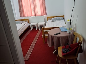 A compact hotel room with two single beds, a small table with a chair, a radiator, and a window with curtains.
