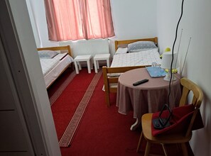 A compact hotel room with two single beds, a small table with a chair, a radiator, and a window with curtains.