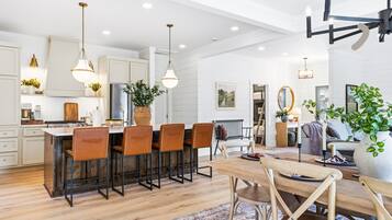 A modern kitchen with bar seating, a dining area with a wooden table, and a living space with a sofa and plants.