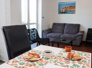 Un coin repas avec une table dressée pour deux personnes, agrémentée d’une nappe à motifs floraux, d’une assiette de nourriture, d’une tasse de café et d’un verre de jus de fruits.