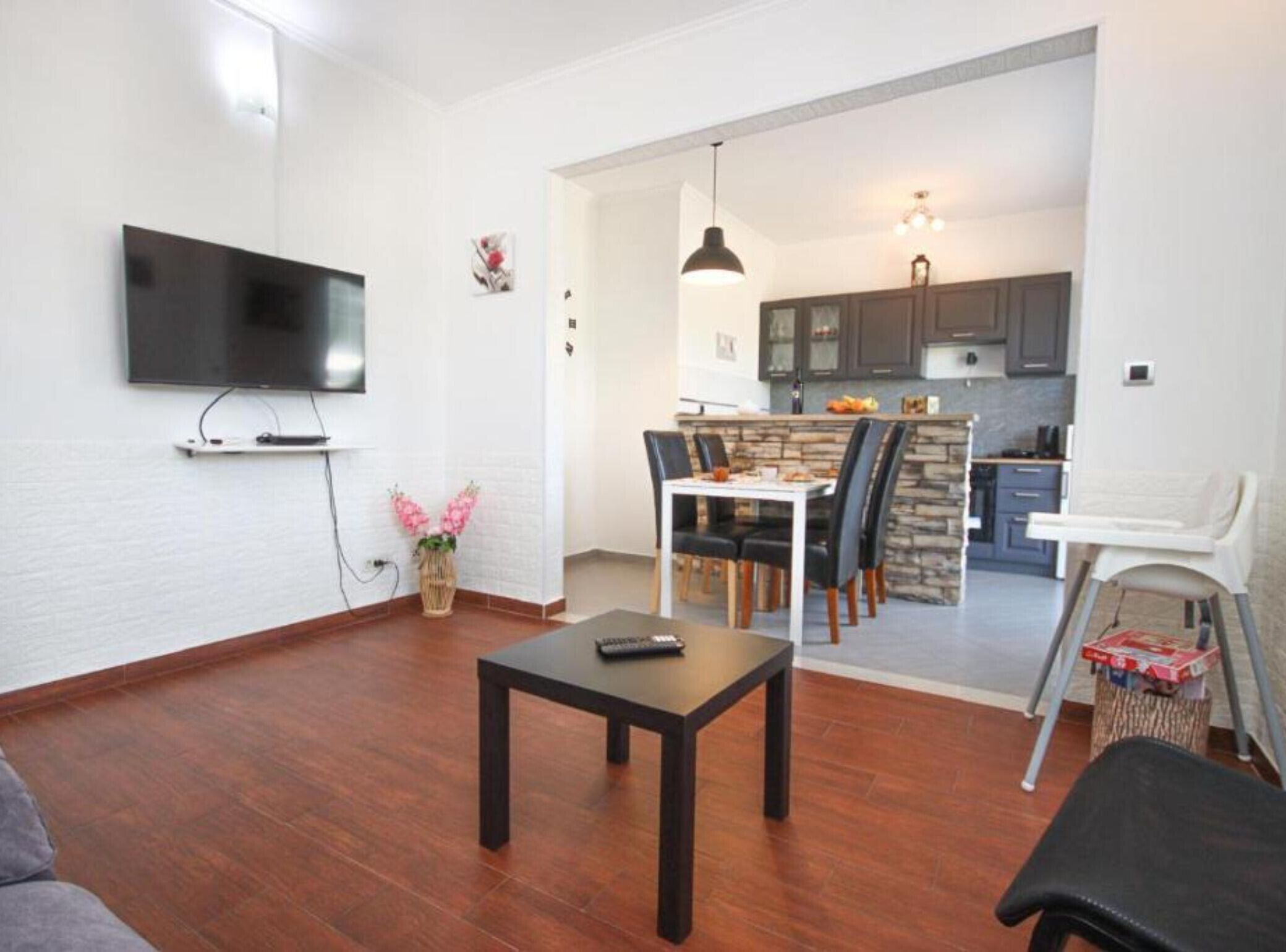 A living room with a TV, a dining table with chairs, a kitchen area with cabinets, and a small dining table with a chair.