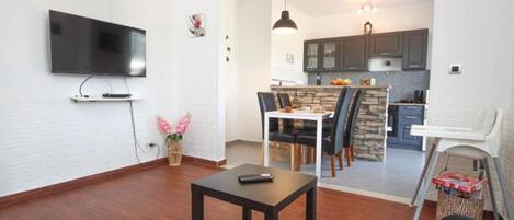 A living room with a TV, a dining table with chairs, a kitchen area with cabinets, and a small dining table with a chair.