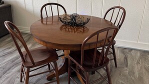 Een ronde houten eettafel met vier stoelen, een geweven decoratiestuk in het midden, en een witte paneelwand.