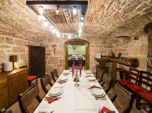 Ein Esszimmer mit Steinsäulen, einem langen Tisch, der für eine Mahlzeit gedeckt ist, Holzsesseln und einem Kruzifix an der Wand.
