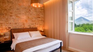 Un dormitorio con pared de piedra, una cama con ropa blanca, vistas a zonas verdes y montañas a través de una ventana, y una lámpara colgante.