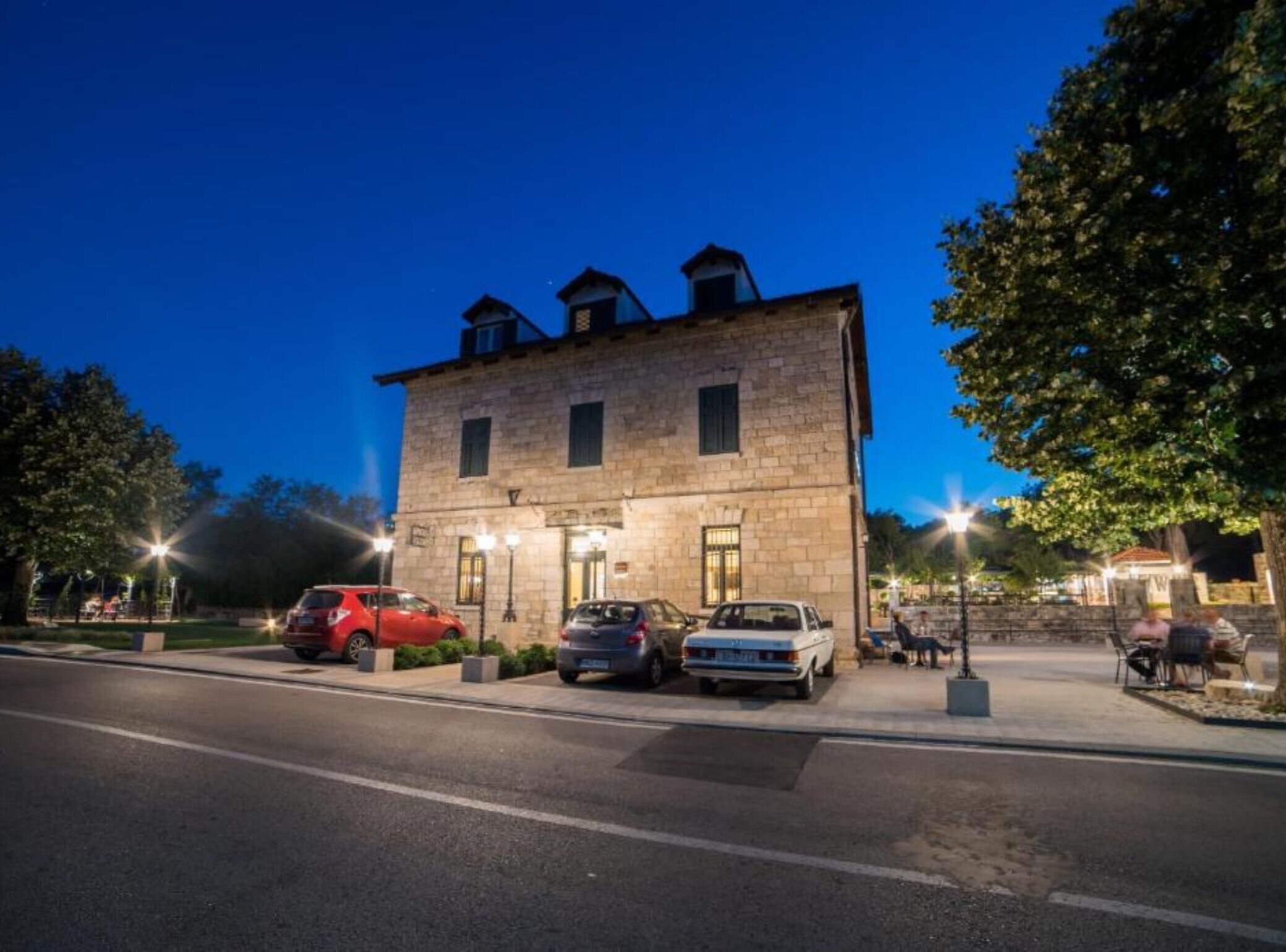 A stone building with a flat roof, two visible windows, and a paved parking area with cars.