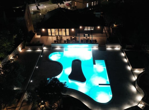 A swimming pool with a unique shape, surrounded by a deck with seating and a building in the background.