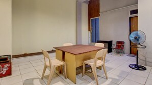 Een eethoek met een houten tafel en vier stoelen, een staande ventilator en een rode stoel in de hoek.