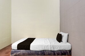 A single bed with a white and black bedspread, a pillow, and a mattress on a wooden floor in a room with plain walls.