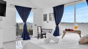 Ein Hotelzimmer mit einem Bett, einem Tisch mit Obst und Getränken, einem Sessel, einem Fernseher und einem Balkon mit Vorhängen.
