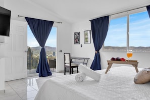 Ein Hotelzimmer mit einem Bett, einem Tisch mit Obst und Getränken, einem Sessel, einem Fernseher und einem Balkon mit Vorhängen.