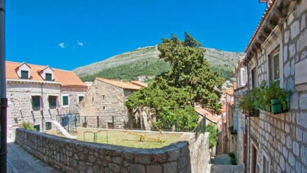 Una estrecha calle de piedra con edificios tradicionales de piedra, un parque infantil y una colina al fondo.
