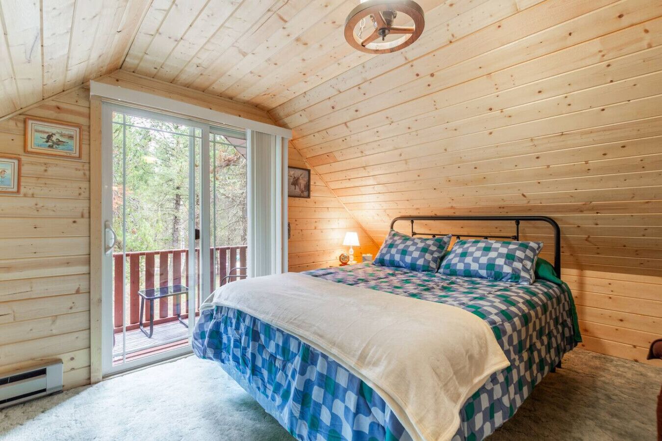 A wooden cabin bedroom with a bed, a nightstand, a lamp, and a view through a sliding glass door to an outdoor deck.
