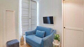 Ein Zimmer mit einem blauen Sofa, einem kleinen Tisch mit einer Pflanze, einem Fernseher an der Wand und einer Schiebetür.