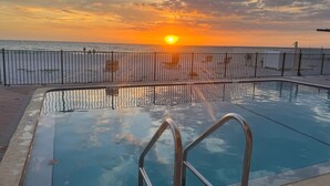 En swimmingpool med gelænder, solnedgang i baggrunden og en strand med et hegn.