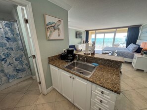 Una cocina con lavamanos, encimera y armarios, un dormitorio con cama y ventana, y un cuadro de una tortuga marina en la pared.