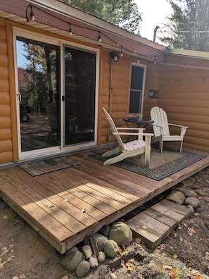 Eine Hütte mit Holzterrasse, zwei Adirondack-Stühlen, einer Glasschiebetür und einem kleinen Tisch.