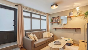 Ein Wohnzimmer mit Sofa, Couchtisch und Fernseher. Es gibt Stufen zu einer Tür, ein Fenster mit Vorhängen und ein Wandregal mit Spiegel und Dekoration.