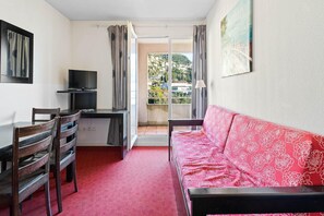 Ein Zimmer mit einem roten, gemusterten Sofa, einem Fernseher auf einem Schrank, einem Esstisch mit Stühlen und Blick durch eine offene Tür.