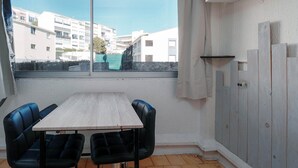 Een kamer met een eettafel, twee stoelen, een raam met uitzicht op gebouwen en een witte muur met een kast.