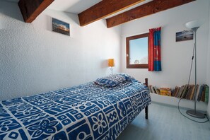 Ein Schlafzimmer mit einem geblümten Bettbezug, einer Nachttischlampe, einem Fenster mit Vorhängen und einem Wandregal mit Büchern.