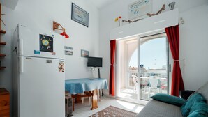 Ett kompakt bostadsutrymme med ett litet kök, en matplats, en TV och en balkong med röda gardiner.
