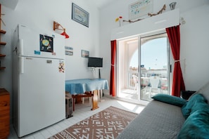 Ein kompakter Wohnbereich mit Küchenzeile, Essplatz, Fernseher und einem Balkon mit roten Vorhängen.