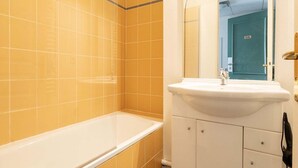 Een badkamer met gele tegels, een wit bad, een wastafel met spiegel en een deur met de tekst 'ELE'.
