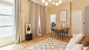 Ein modernes Wohnzimmer mit Essbereich, großen Fenstern mit Vorhängen, einer Kristallleuchtre und einem gerahmten Kunstwerk an der Wand.