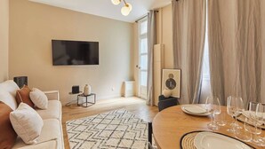 Ein modernes Wohnzimmer mit einer Couch, einem Fernseher, einem Esstisch gedeckt mit Gläsern und einem großen Fenster mit Vorhängen.