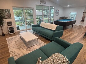 Un salon avec une table de billard, un canapé vert, une table basse blanche et une grande fenêtre donnant sur une terrasse.