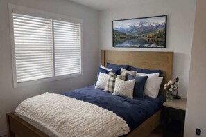 A bedroom with a bed, a window with blinds, a headboard, a painting of a landscape, and a nightstand with a vase of flowers.