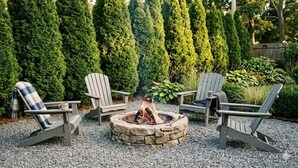 A stone fire pit with four Adirondack chairs, surrounded by gravel and tall evergreen trees. - Home 2 mins. from Shinnecock Golf town SH beaches (Southampton)