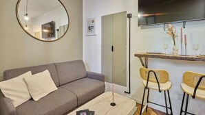 Un espace de vie comprenant un canapé, un miroir rond, une table à manger avec des chaises, et un grand téléviseur à écran plat fixé au mur au-dessus de la table.