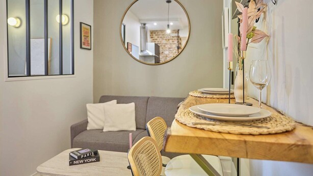 Un coin repas avec un miroir rond, une table dressée avec des assiettes et un verre à vin, un canapé gris et un petit livre posé sur une table d’appoint.