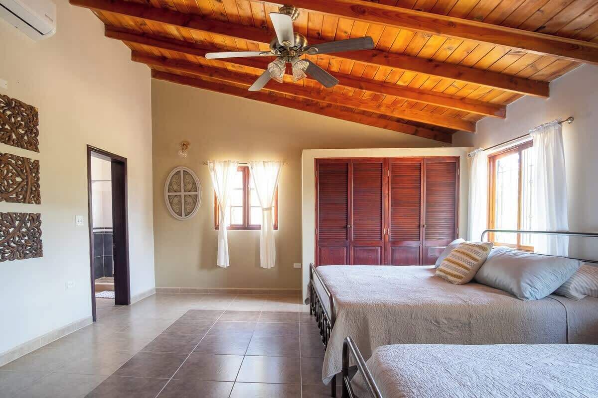 A bedroom with a bed, a ceiling fan, wooden beams, a window, and a door.