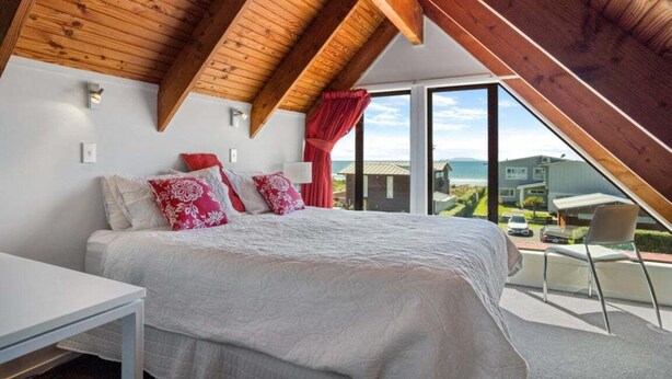 Ein Schlafzimmer mit schrägverdeckter Decke, einem Bett mit weißer Bettwäsche und roten Kissen, einem Nachttisch und Blick ins Freie.