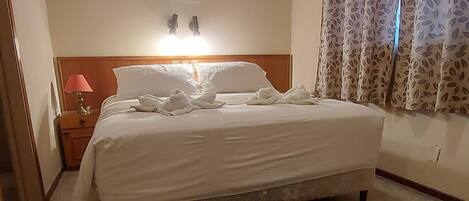 Una habitación de hotel con cama, mesita de noche, lámpara, un cuadro en la pared y ventana con cortinas estampadas.