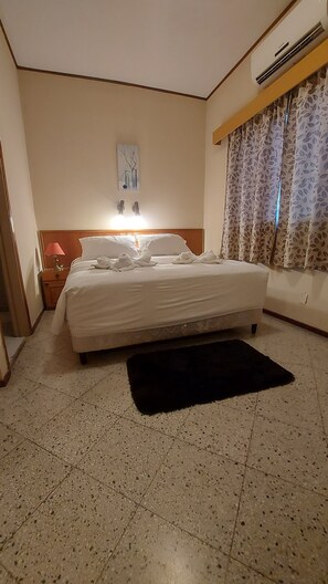 A hotel room with a bed, a nightstand, a lamp, a painting on the wall, and a window with patterned curtains. - Paranainn Hotel Asuncion (Asunción)