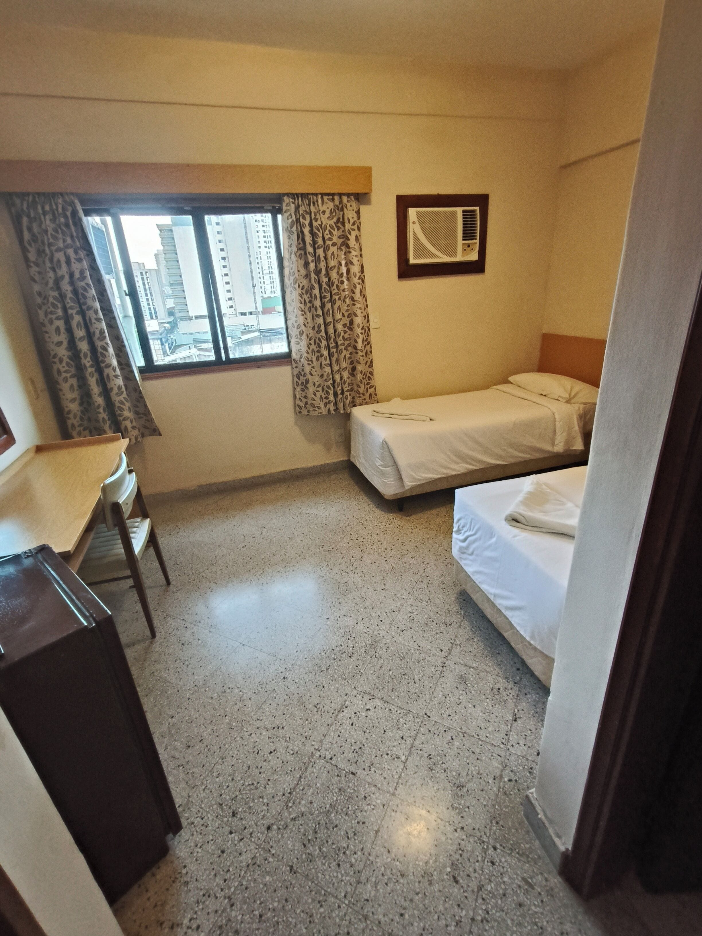 A small hotel room with a bed, a desk, and a window with curtains.