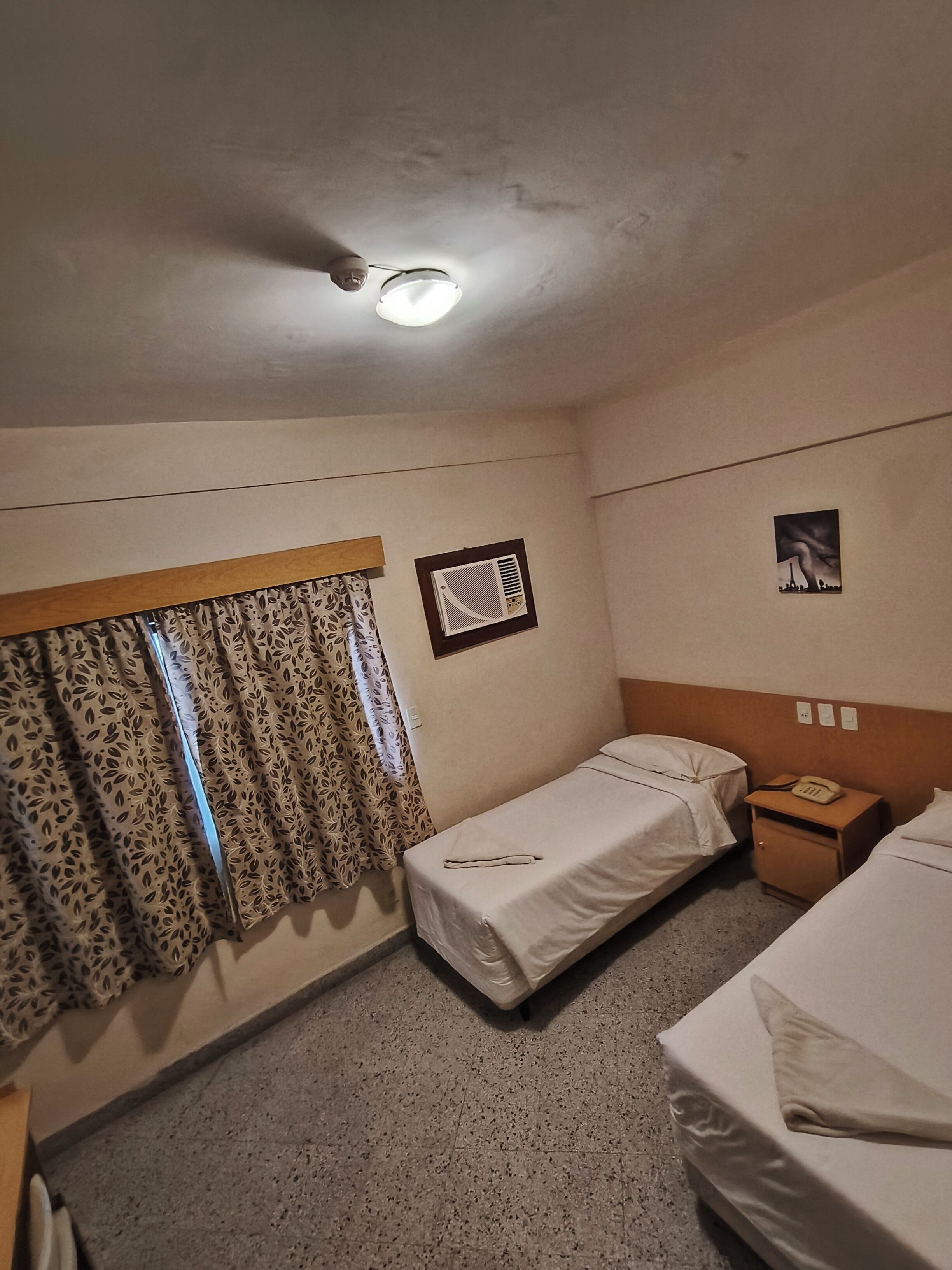 Una camera d'albergo con due letti singoli, un comodino con il telefono, un termosifone a muro e una finestra con tende a motivi.